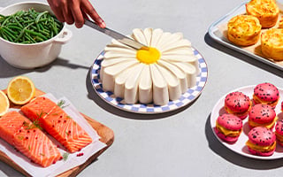 Table dress&eacute;e avec un dessert en forme de marguerite en cours de d&eacute;coupe, accompagn&eacute; de haricots verts, de pav&eacute;s de saumon, de pommes de terre gratin&eacute;es et de petits desserts color&eacute;s.