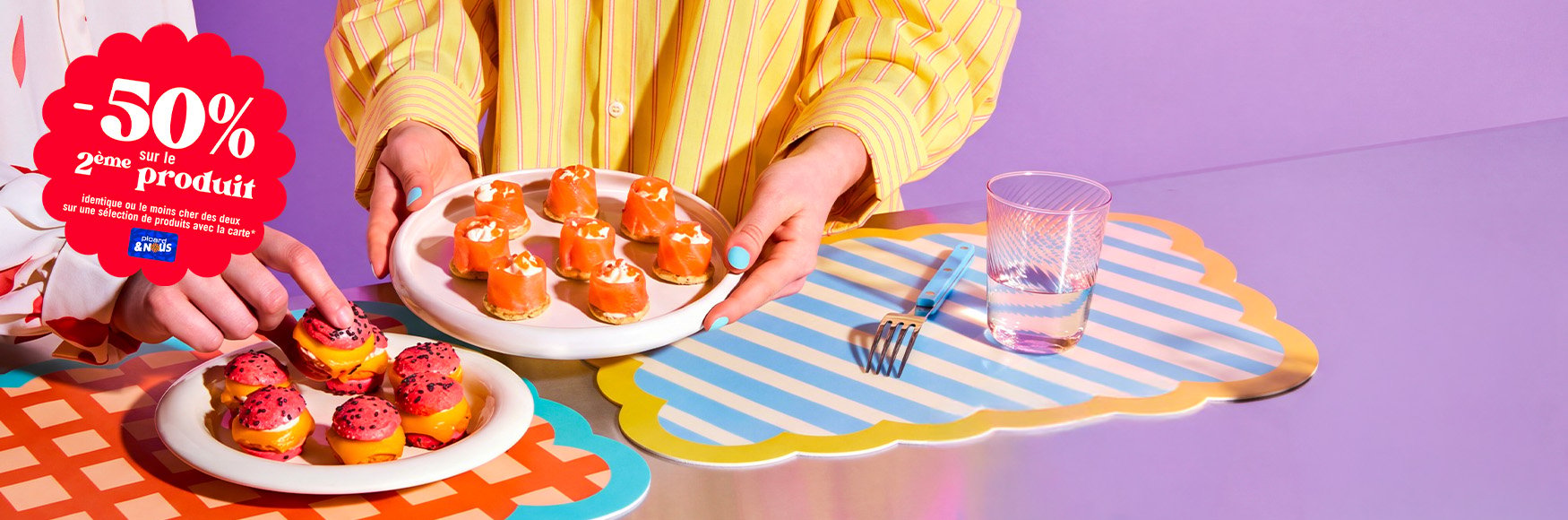  Deux mains tenant des assiettes de bouch&eacute;es ap&eacute;ritives color&eacute;es pos&eacute;es sur une table, avec une offre promotionnelle indiquant &ldquo;-50 % sur le deuxi&egrave;me produit&rdquo;. `